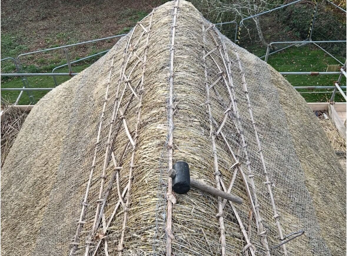 A thatched ridge in progress, hazel spars crossing the apex with a wooden mallet resting on the straw