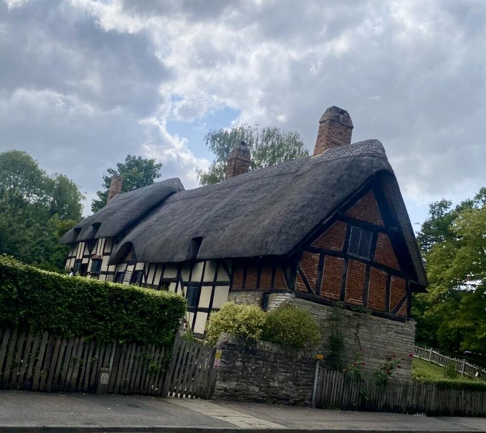 A thatched half-timbered cottage with brick infill panels and multiple chimneys behind a stone wall and wooden fence